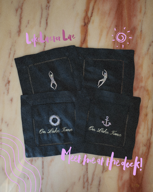 Four black embroidered fabric coasters or small mats arranged on a light wood surface. Each piece has a stitched border and a white nautical-style emblem: one figure, one anchor, one ring, and one swimmer, with the text 'On Lake Tahoe' on two pieces.