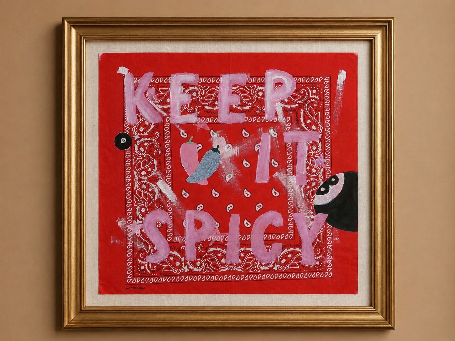 Framed wall art featuring a red background with the words 'KEEP IT SPICY' in light pink letters, overlaid with white paisley bandana patterns and small abstract blue and black details, shown in a gold frame against a beige wall.