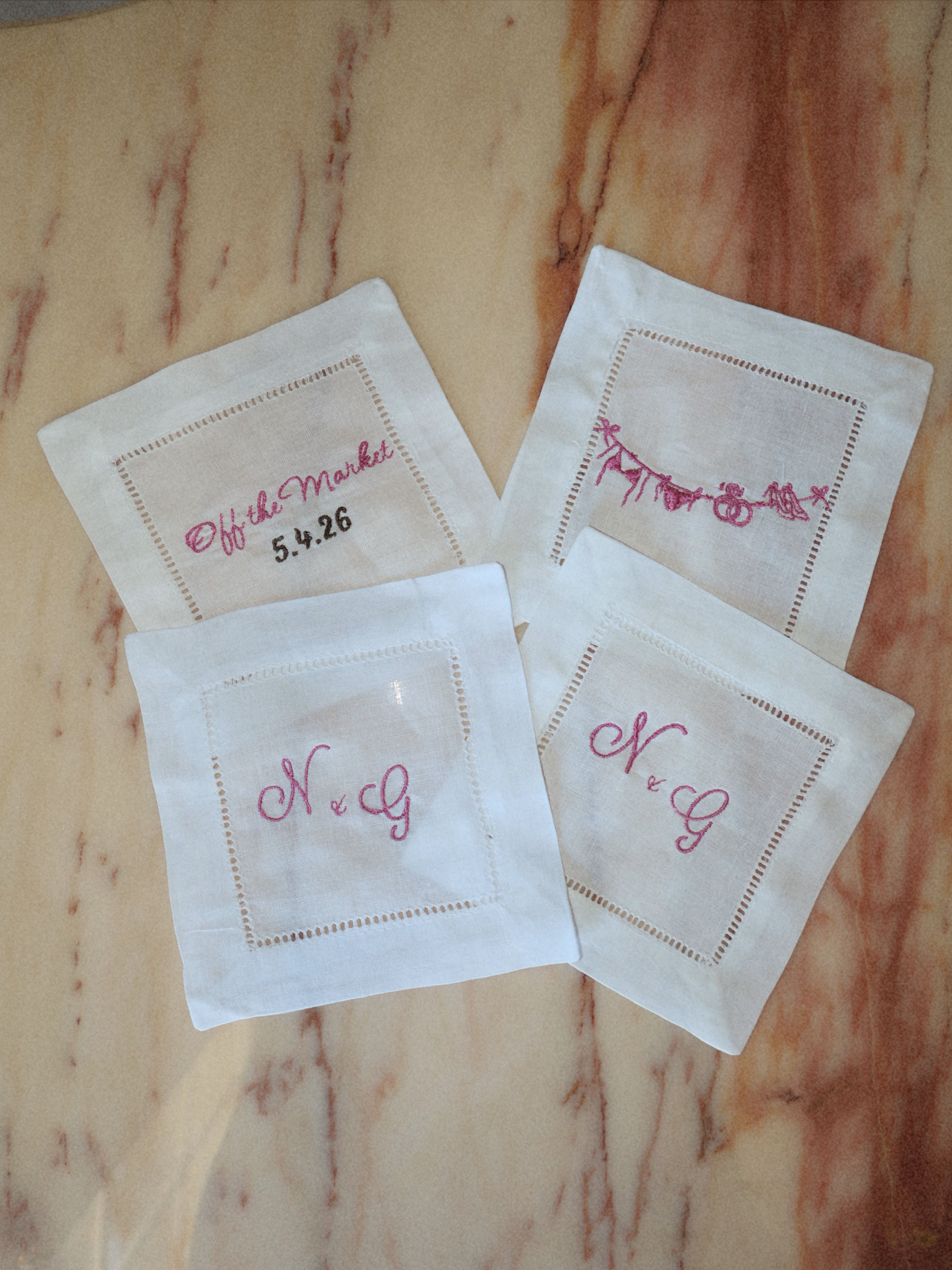 Four white embroidered cocktail napkins with pink script monogram names and dates, arranged on a light brown marble surface.