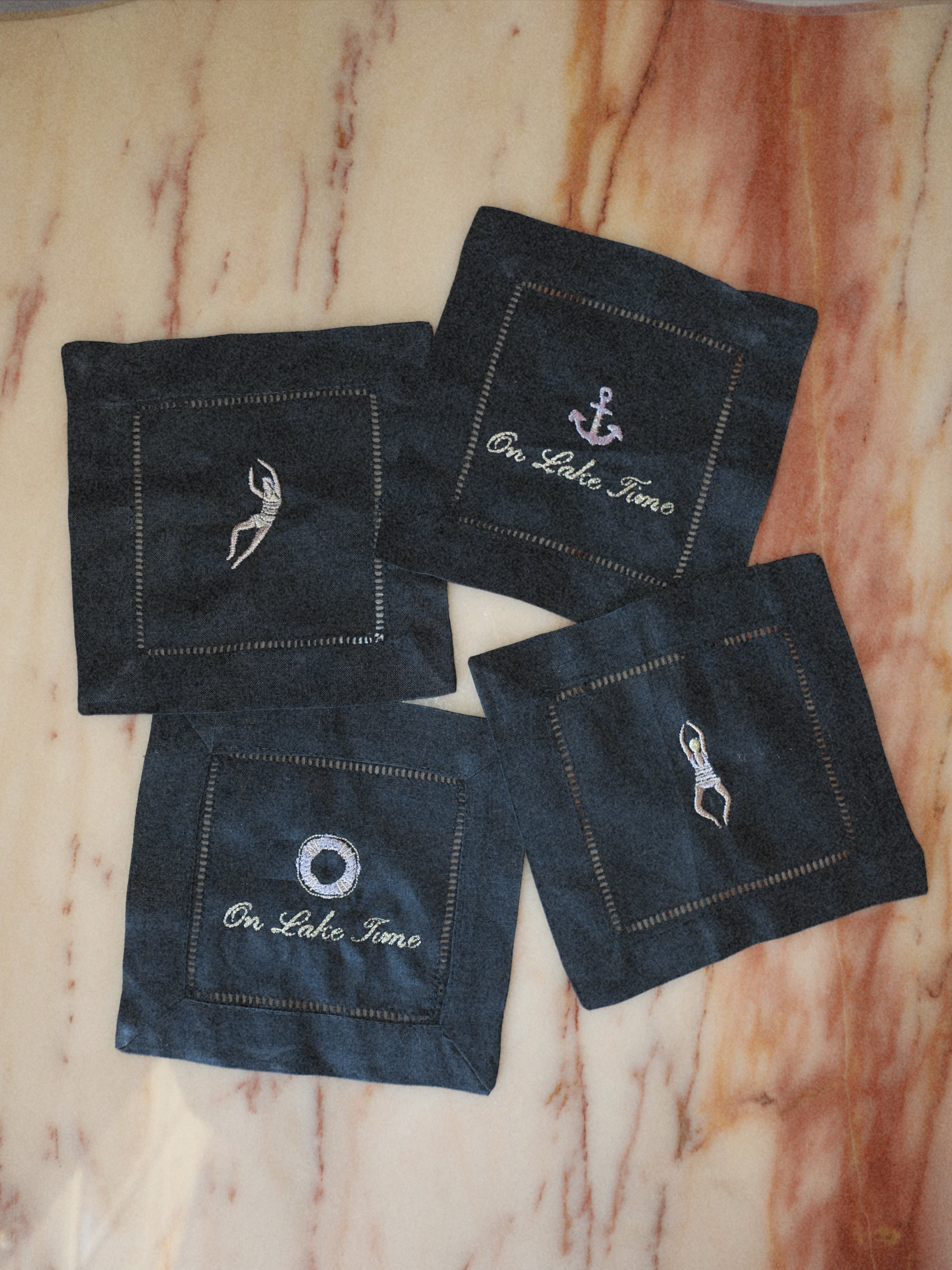 Four dark blue denim-like square fabric coasters with white embroidered nautical designs and text, arranged on a beige marble-patterned surface.