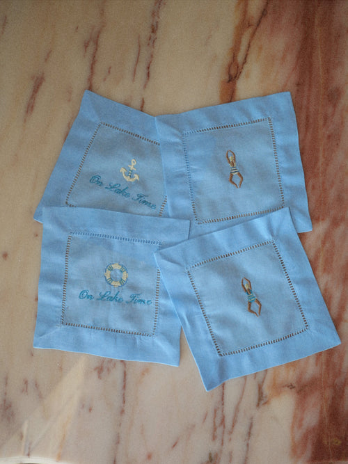 Four light blue embroidered square napkins arranged on a brown marble-patterned surface. Each napkin has a stitched border and a small central embroidery in blue, gold, and white.